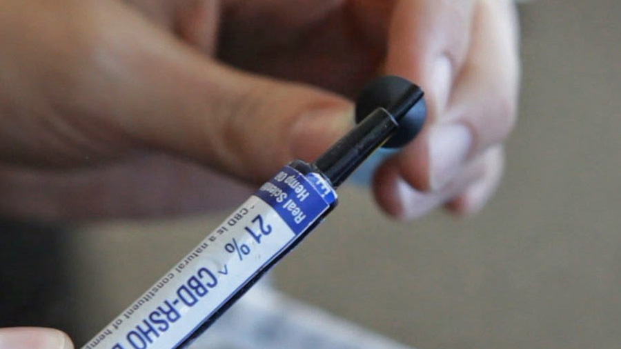 Mãos de uma pessoa manuseando êmbolo e seringa com rótulo onde está escrito em azul “21% CBD-RSHO Blue”. Importação.