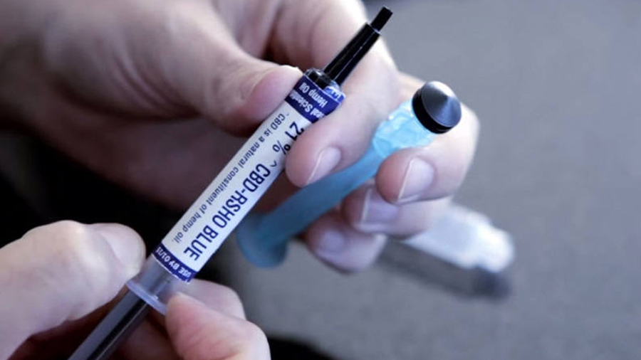 Fotografia de uma seringa de canabidiol com o texto “21% CBD-RSHO Blue” em azul sobre fundo branco em seu rótulo, sendo segurada por uma mão que também segura um êmbolo entre os dedos e outra mão que segura sua base. Foto: Anvisa.