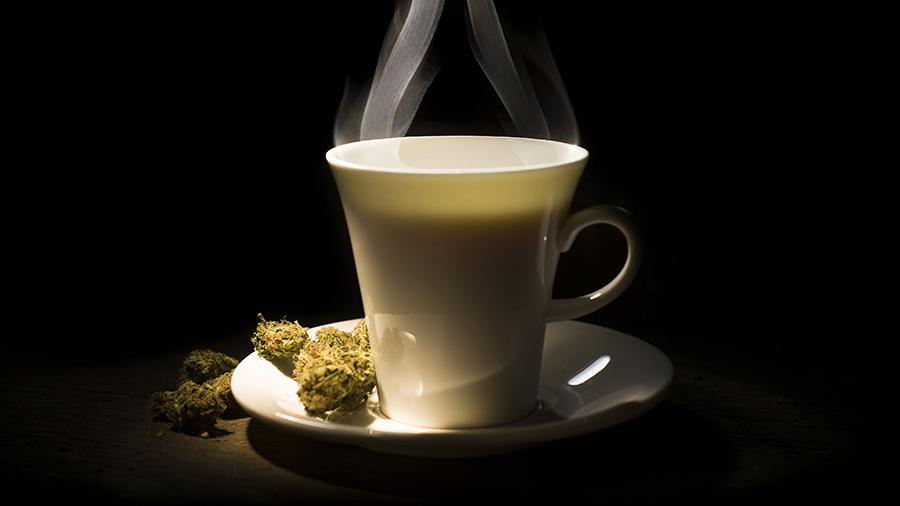Uma xícara branca sob um feixe de luz diagonal com fundo escuro, de onde sai o vapor do café, junto a algumas flores de maconha sobre um pires.