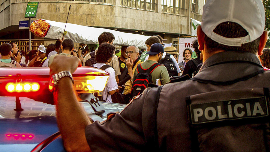 Fotografia que mostra a parte de cima das costas de um policial militar fardado, usando boné branco e com uma das mãos apoiadas no giroscópio de uma viatura (no primeiro plano), enquanto observa transeuntes durante a Marcha da Maconha no Rio de Janeiro. PM.