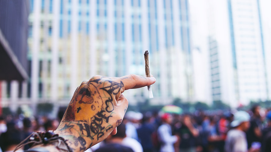 Fotografia de uma mão tatuada segurando um baseado. Foto: Diogo Vieira | Smoke Buddies (Cobertura Marcha da Maconha de SP, 2017).