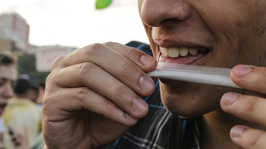 Fotografia em plano fechado que mostra partes das mãos e do rosto (do nariz para baixo) de uma pessoa que está fechando um cigarro de maconha. SUS.