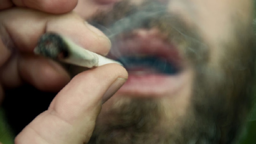 Uma pessoa fumando um baseado que aparece em primeiro plano sendo segurado pelos dedos polegar e indicador e ao fundo a boca aberta expelindo fumaça. Legalização.
