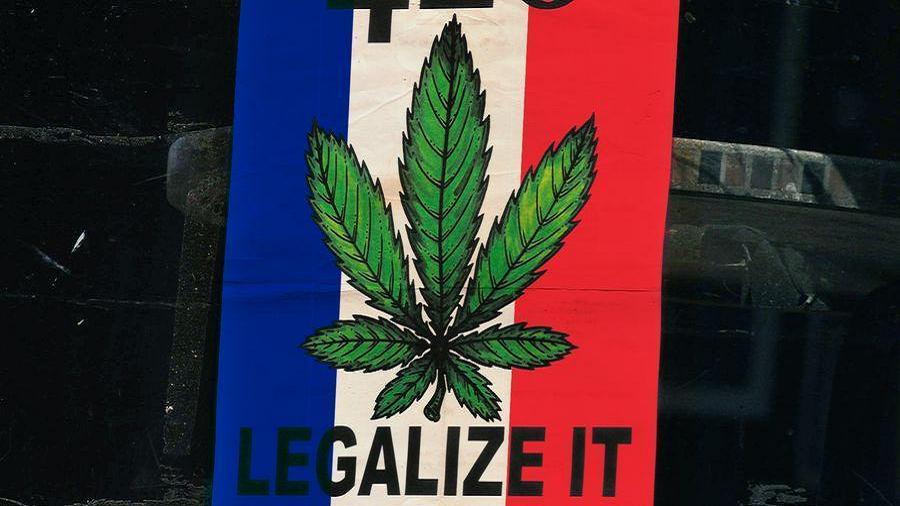 Fotografia de uma placa onde está impressa o desenho de uma folha de cannabis (maconha) e a escrita “Legalize it” sobre a bandeira da França sob  e um fundo escuro.