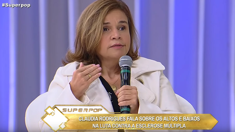 Atriz com um microfone na mão e uma legenda do programa Superpop com o texto "Claudia Rodrigues fala sobre os altos e baixos na luta contra a esclerose múltipla".