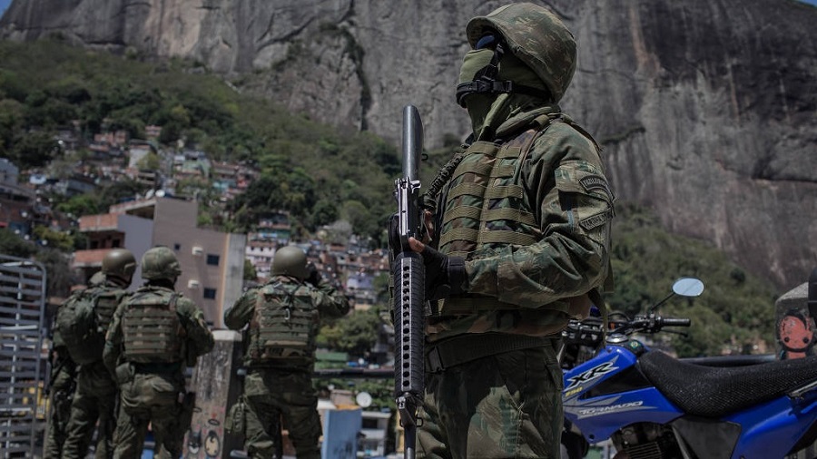 Agente militar das Forças Armadas segurando um fuzil apontado para baixo e olhando para cima, e ao fundo outros militares observam o morro, durante intervenção realizada na favela da Rocinha.