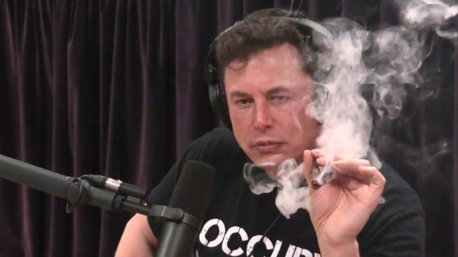 Elon Musk usando fones de ouvido tipo circumaural, atrás de um microfone e segurando um baseado aceso entre fumaça. Tesla.
