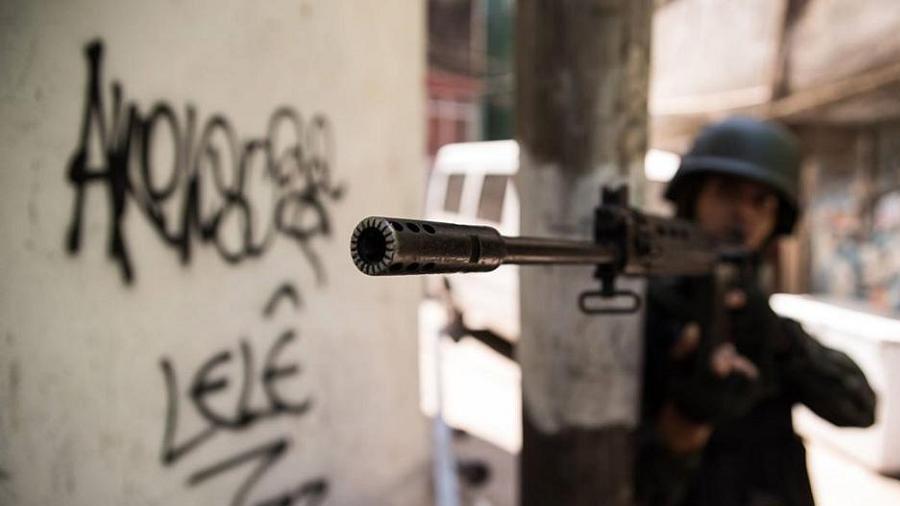 Um fuzil sendo apontado por um agente do exército que se apoia em um poste, com foco no quebra-chamas, nas ruas do Rio de Janeiro. Drogas.