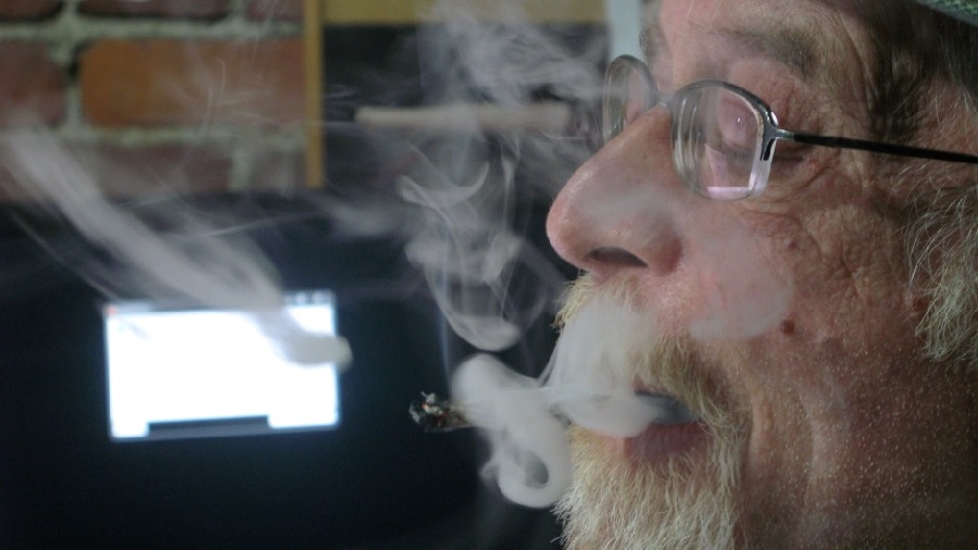 Fotografia em perfil da face de um homem idoso, usando óculos e barba, que expele uma fumaça densa, em ambiente interno, e um fundo desfocado.
