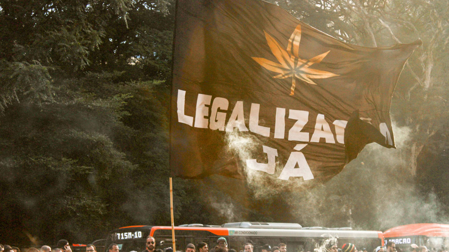 Uma bandeira preta enorme com o desenho de uma folha de maconha em cor dourada e escrito em branco “Legalização Já”, hasteada pelos manifestantes durante a Marcha da Maconha de São Paulo, em 2017. Liberdade.