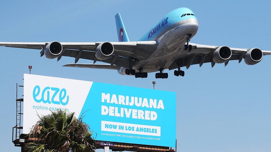 Um avião sobrevoando um outdoor nas cores azul claro e branco com o texto "Eaze. Marijuana Delivered. Now in Los Angeles". Maconha.