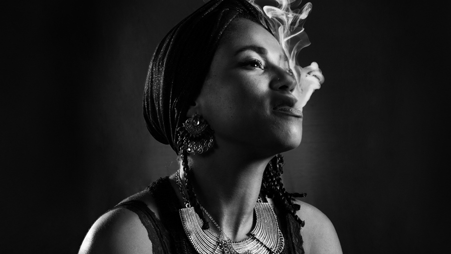 Fotografia em preto e branco, primeiro plano e meio perfil de uma mulher negra vestida com adornos africanos e olhando para cima, enquanto expele delicadamente uma fumaça densa. 2019.