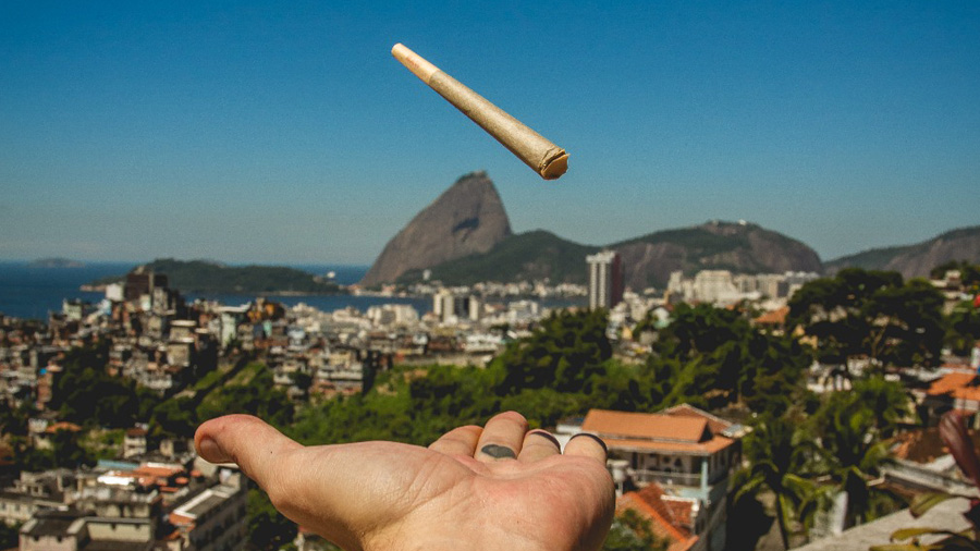 Fotografia da mão de uma pessoa (que está atrás da câmera) lançando para cima um baseado e ao fundo vemos parte da cidade do Rio de Janeiro, o Pão de Açúcar e o céu em um degrade de azul. Viagem.