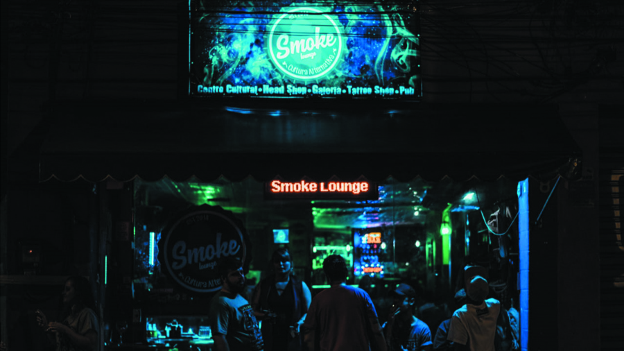 Fotografia colorida frontal, tirada à noite, que registra o movimento de pessoas em frente ao centro cultural que tem seu nome "Smoke Lounge" em letreiro luminoso na entrada. Head shops.