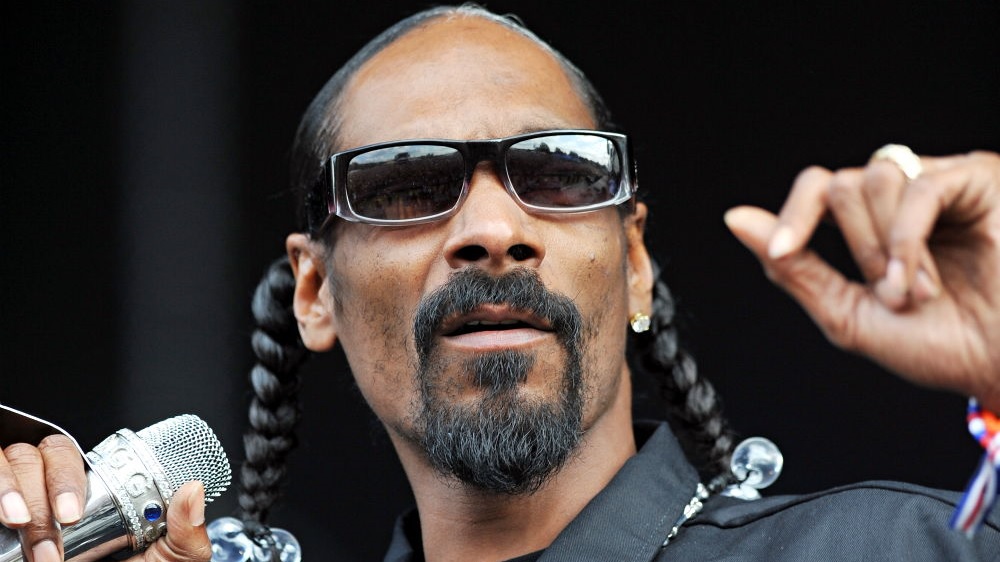 Fotografia frontal em primeiríssimo plano de Snoop Dogg usando óculos escuros e segurando um microfone prateado.