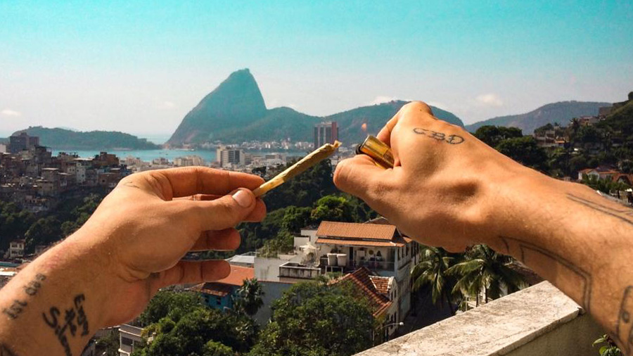 Fotografia dos braços tatuados de uma pessoa que está atrás da câmera e segurando na mão esquerda um baseado e na mão direita um isqueiro dourado. Ao fundo vemos árvores, casas, prédios, parte da enseada de Botafogo, o Pão de Açúcar e o céu em azul degradê, no Rio de Janeiro. Smoke Buddies.