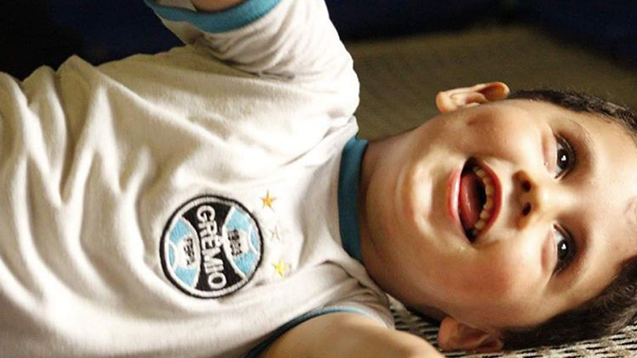 Fotografia em meio primeiro plano do pequeno Kauê deitado e sorridente, vestindo roupa branca, com o brasão do Grêmio (nas cores preta e azul), com detalhes nas mangas e gola na cor azul. Autismo.