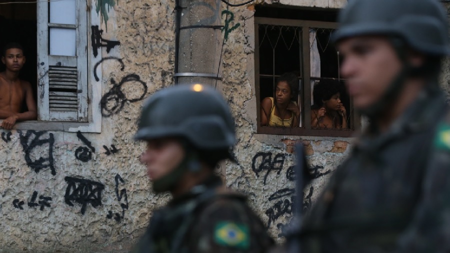 Fotografia em vista lateral diagonal de dois agentes do exército brasileiro (usando capacete) lado a lado, no primeiro plano desfocado, e ao fundo moradores observam apreensivos pelas janelas, no bairro de Anchieta, zona norte do Rio de Janeiro, durante intervenção militar em 2018.