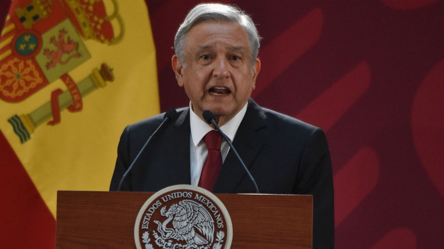 Fotografia em primeiro plano de Andrés Manuel López Obrador, falando atrás de dois microfones e um púlpito de madeira escura, que tem o brasão nacional do México em branco; ao fundo pode-se ver parte da bandeira espanhola arriada e uma parede em tons de vermelho.