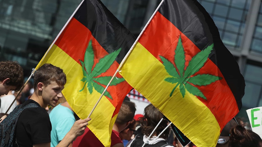 Fotografia de duas bandeiras da Alemanha com o desenho da folha da maconha ao centro, sendo carregadas na diagonal por jovens que participam da Hanfparade (Parada da Maconha).
