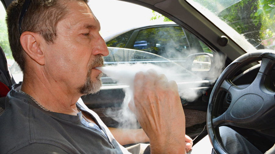 Fotografia em primeiro plano e perfil de um homem sentado ao volante, enquanto fuma um baseado que segura próximo à boca ao expelir uma fumaça densa; ao fundo pode-se ver parte da lateral de outro carro próximo. Colorado.