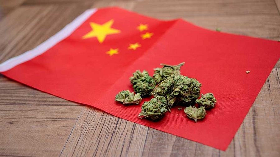 Fotografia em vista superior de uma porção de flores secas de maconha sobre uma pequena bandeira da China que está sobre uma superfície de madeira. Ações.