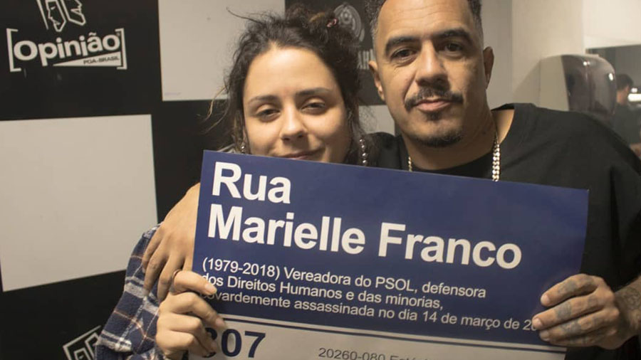 Fotografia em primeiro plano de Nah Brisa segurando, junto com Marcelo D2, uma placa (azul e branco) da rua em homenagem a Marielle Franco. Minas.