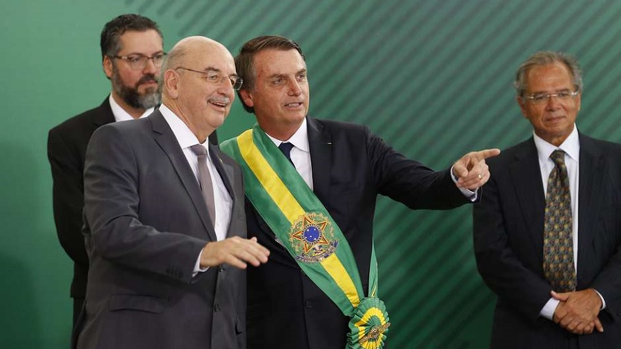 Fotografia de Osmar Terra sorridente, junto a Jair Bolsonaro que está usando a faixa presidencial e apontando para algo que os dois observam; ao fundo, estão os ministros Paulo Guedes, à direita, e Ernesto Araújo, à esquerda e atrás de Osmar, próximos de uma parede verde.