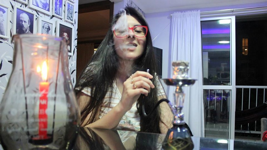 Fotografia da deputada Ana Caroline Campagnolo sentada atrás de uma mesa de vidro redonda, enquanto segura a mangueira de um narguilé que está sobre a mesa e expele fumaça; na parte esquerda da foto, pode-se ver uma vela vermelha acesa, no primeiro plano.