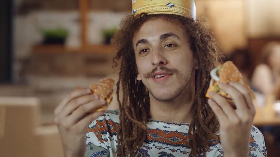 Frame do vídeo do Burger King, onde vemos o personagem usando dreads e uma coroa e segurando dois pedaços de lanche, com um fundo desfocado.