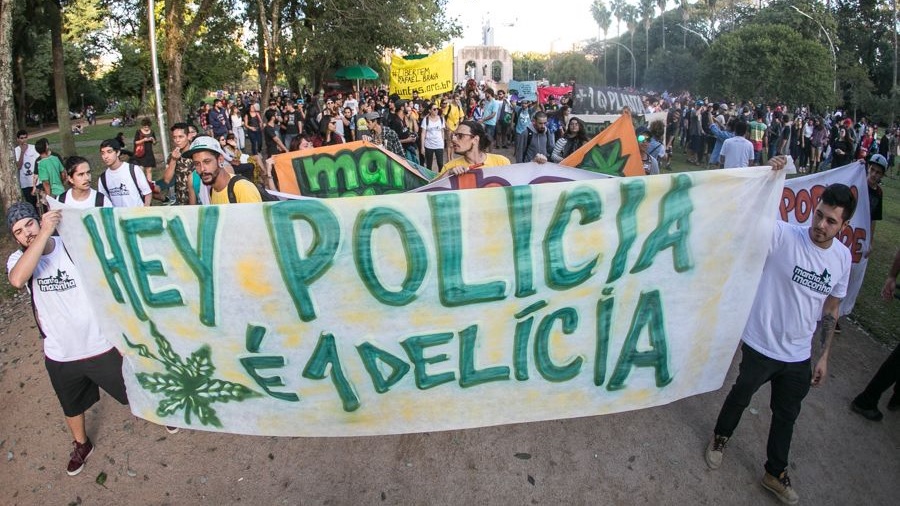 Fotografia em vista superior que mostra dois manifestantes segurando uma faixa branca com o texto "Hey polícia [o desenho de uma folha de maconha] é 1 delícia" em verde, à frente de uma multidão que segue por um parque, durante a Marcha da Maconha de Porto Alegre, em 2017.