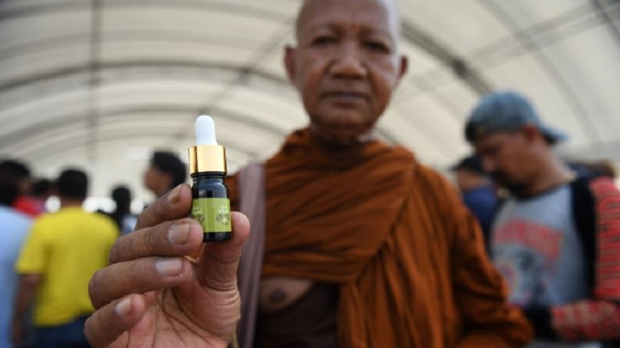 Fotografia em primeiro plano do monge budista segurando um frasco de óleo de cannabis diante da câmera; ao fundo pode-se ver outras pessoas, parcialmente nítidas, que participam do festival. Tailândia.