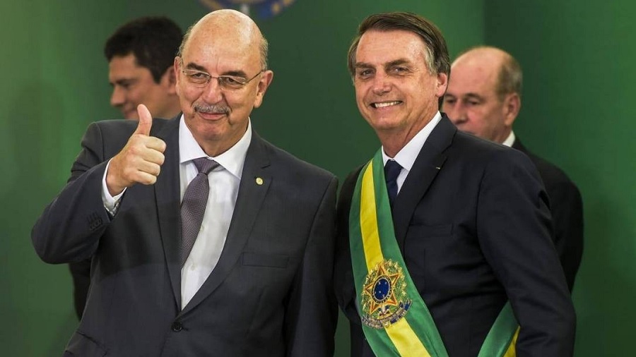 Fotografia em primeiro plano de Osmar Terra, fazendo sinal de positivo, ao lado de Jair Bolsonaro, usando a faixa presidencial, ambos sorridentes; ao fundo, podemos ver as cabeças dos ministros Sergio Moro (atrás de Terra) e Fernando Azevedo (atrás de Bolsonaro). Drogas.