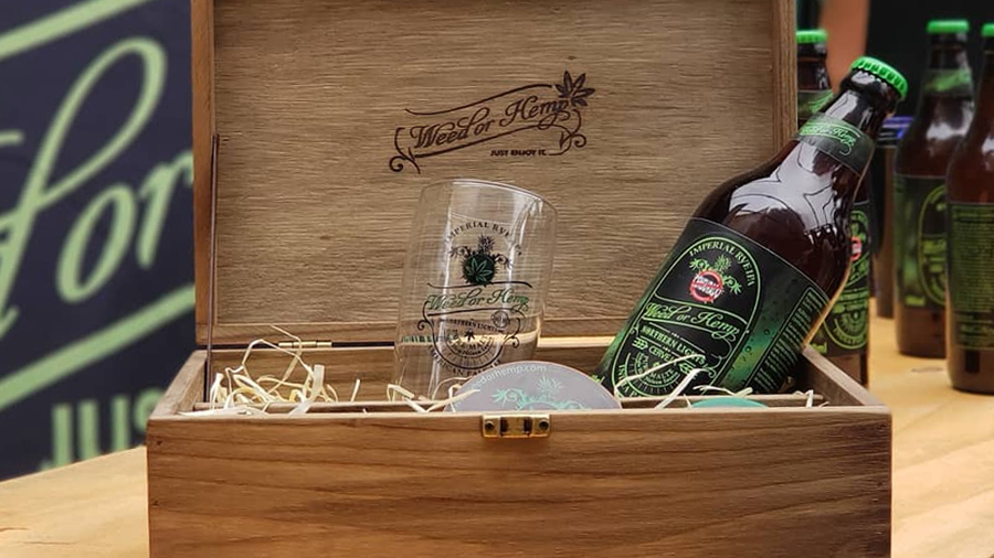 Fotografia em plano fechado de uma caixa promocional da Weed or Hemp, feita de madeira, com um copo e uma garrafa da cerveja Northern Lights, no primeiro plano, e, no segundo, parte direita da foto, algumas garrafas dispostas no balcão.
