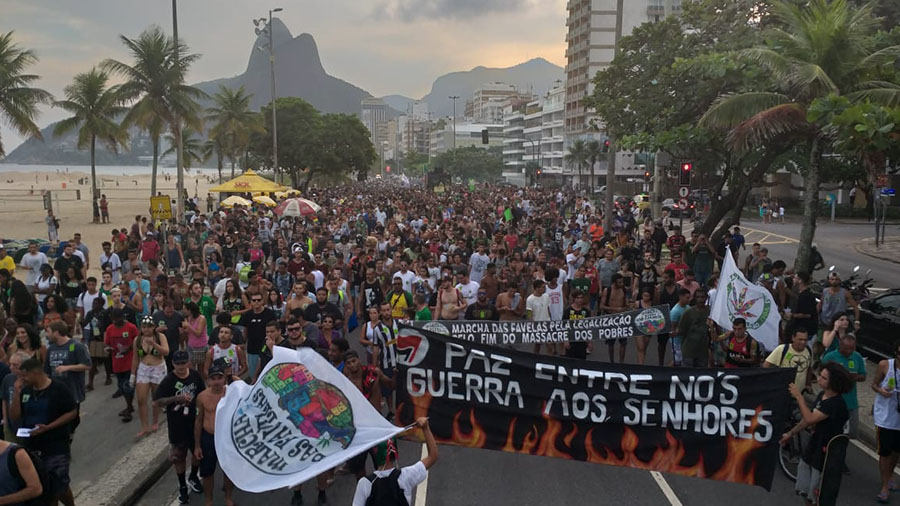 Fotografia mostra, do ponto de vista superior, milhares de pessoas durante a Marcha da Maconha no Rio de Janeiro; na parte inferior direita da foto, logo à frente dos manifestantes pode-se ver uma faixa preta com o texto em branco "Paz entre nós - Guerra aos senhores" e o desenho de chamas na parte inferior.