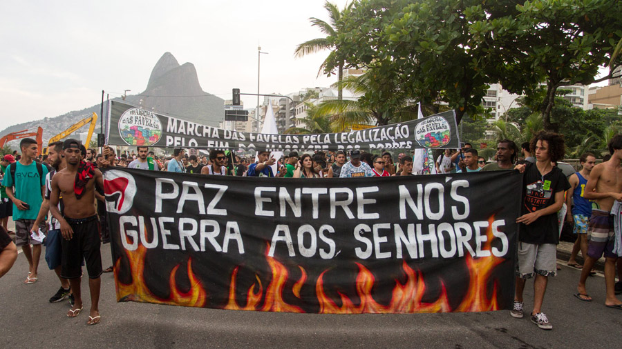 Fotografia frontal da Marcha da Maconha, realizada no Rio de Janeiro. Nela há grandes faixas onde se lê “Paz entre nós guerra aos senhores” e "Marcha das favelas pela legalização, pelo fim do massacre aos pobres". Paz.