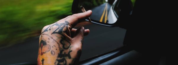 Fotografia tirada do interior de um carro que mostra uma mão tatuada segurando um baseado (cigarro de maconha) sobre o vidro abaixado, o retrovisor mostrando a pista de faixa contínua e uma paisagem verde desfocada (em movimento), ao fundo. Imagem: Diogo Vieira.