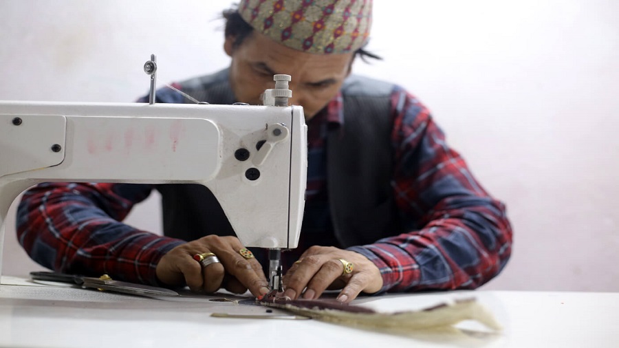 Fotografia mostra um senhor atrás de uma máquina de costura branca. Ele veste um chapéu estampado, uma camisa xadrez e usa anéis nos dedos, que costuram uma peça de roupa. Foto: Nepali Times.