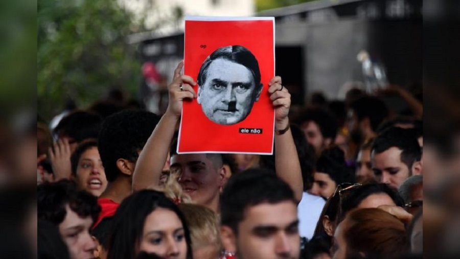 Fotografia retrata uma aglomeração de rostos desfocados e, ao centro, uma pessoa com os braços erguidos e um cartaz, com fundo vermelho, uma colagem com metade do rosto de Bolsonaro, metade de Hitler, e a frase "ele não".  Foto: Euromade.
