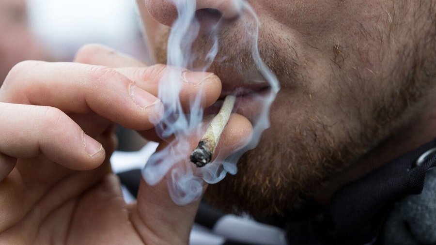 Fotografia em plano fechado que mostra a boca e mão de uma pessoa que fuma um baseado de onde sai fumaça. Drogas.