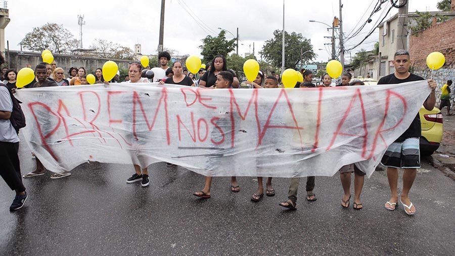 Fotografia que mostra manifestantes carregando balões amarelos e uma faixa branca que traz escrito em vermelho "Parem de nos matar", durante o velório da menina Ágatha Félix. Witzel.