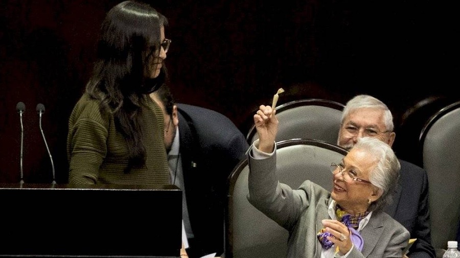 Fotografia em primeiro plano de Olga Sánchez Cordero sorridente, sentada e segurando um cigarro enrolado de maconha, enquanto olha para deputada Lucía Riojas, que está em pé ao seu lado, na Câmara de Deputados do México.
