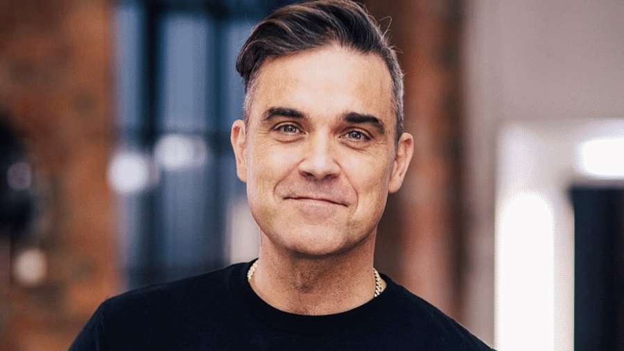 Recorte de uma foto de Robbie Williams que mostra o cantor dos ombros pra cima, vestindo camiseta de cor azul-escuro e com um sorriso fechado.