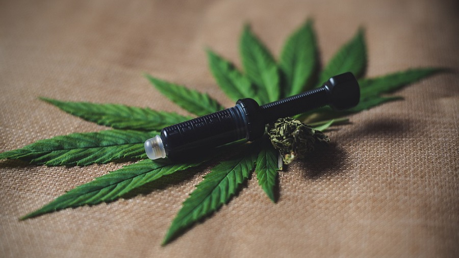 Fotografia que mostra uma seringa preta junto a um pequeno bud seco e duas folhas verdinhas de maconha (cannabis), sobre um pedaço de tecido que parece ser feito de algodão cru. Foto: Pixabay.