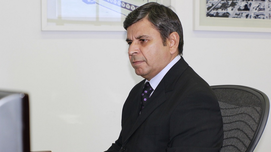 Fotografia em primeiro plano e meio perfil do Juiz Federal Walter Nunes da Silva Júnior, sentado em uma poltrona com encosto de tela e vestindo um paletó preto; ao fundo, uma parede branca e partes de dois quadros com molduras brancas.
