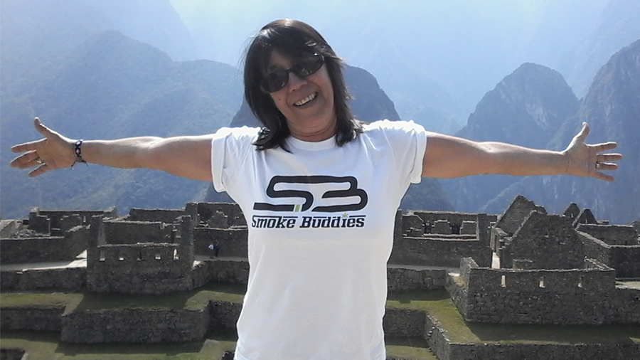 #PraCegoVer: Em destaque, fotografia da Dinda Márcia Scartezini de braços abertos, vestindo camiseta branca com a marca Smoke Buddies, em Machu-Picchu, no Peru. Fotografia: Arquivo Pessoal