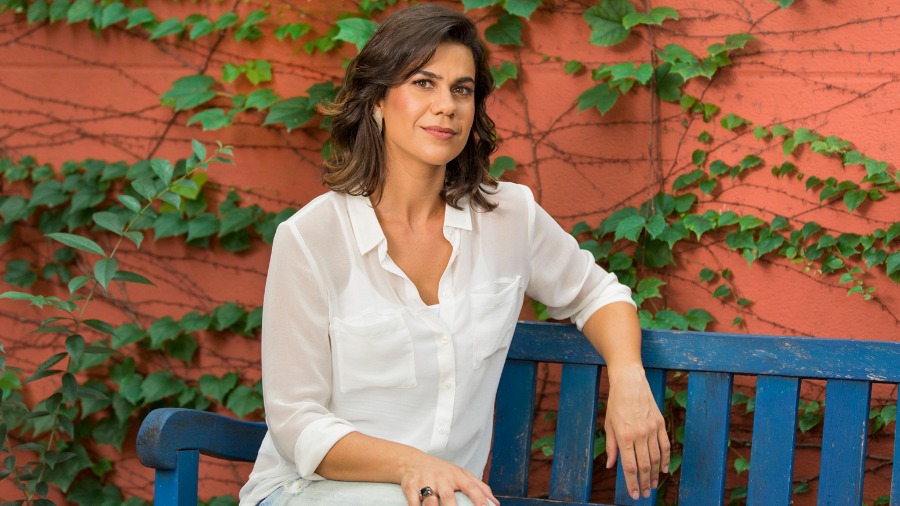 Fotografia em primeiro plano da empresária Viviane Sedola, vestindo uma camisa branca e sentada em um banco azul; ao fundo, uma parede de cor terracota com plantas trepadeiras. Female 50.