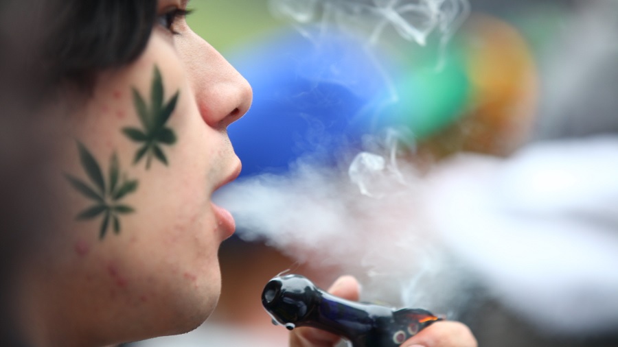 Fotografia que mostra uma pessoa jovem, em perfil e close-up, expelindo fumaça e segurando um pipe de vidro de cor azul-escuro, e um fundo desfocado e colorido; detalhe para dois desenhos de folha de maconha, em verde-escuro, na bochecha. Foto: GoToVan | Flickr.