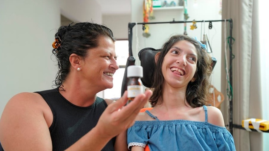 Fotografia em primeiro plano de Regiane, que segura um frasco de medicamento à base de cannabis, enquanto olha para a filha, Júlia, à sua esquerda, ambas sorridentes. Pacientes.
