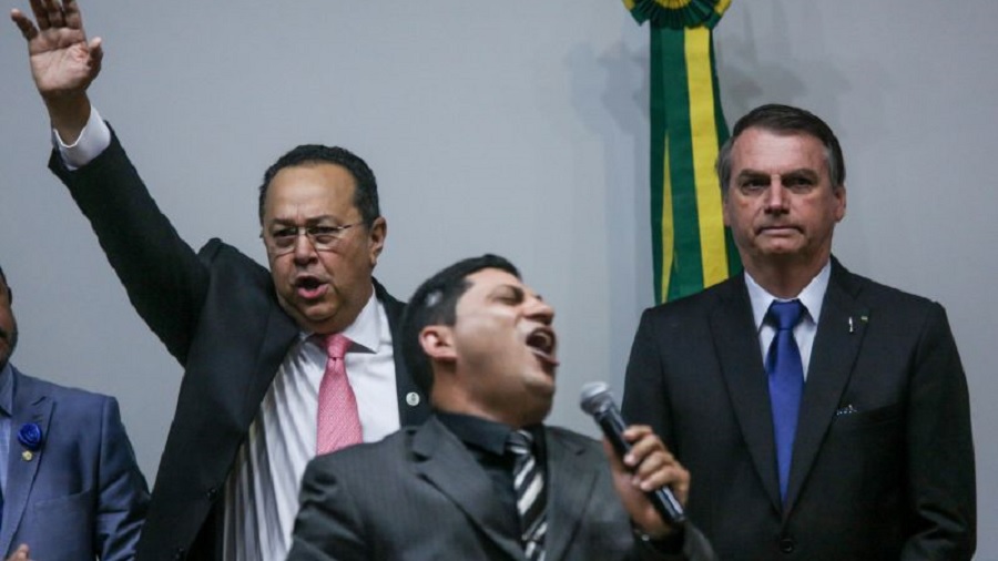 Fotografia em primeiro plano de um pastor, com o corpo inclinado para trás, enquanto canta ao microfone, e, logo atrás, Silas Câmara, com a mão erguida e cantando, e Jair Bolsonaro, durante culto na Câmara dos Deputados.